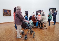 Bildbeschreibung: Eine Gruppe betrachtet Gemälde in einem Ausstellungsraum. Die Gruppe besteht mehrheitlich aus älteren Personen. Eine Frau im Vordergrund stützt sich auf einen Rollator, andere haben sich auf eine Sitzinsel und Klapphocker gesetzt.