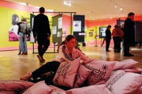 Bildbeschreibung: Eine junge Frau sitzt in einem Stapel aus bedruckten Kissen und liest einen Text, der auf eines der Kissen gedruckt ist. Dahinter erstreckt sich die Ausstellung, in der mehrere Personen die Plakate und Texte betrachten.