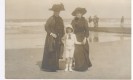 Bildbeschreibung: Die historische Fotografie zeigt zwei in schwarzen Kleidern gekleidete Damen mit gro&szlig;en H&uuml;ten und ein wei&szlig; gekleidetes M&auml;dchen mit Hut am Strand. Im Hintergrund ist das Meer zu sehen. Das Foto ist um 1900 entstanden und daher schwarz-wei&szlig;.