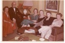 Bildbeschreibung: Die Farbfotografie der 1950er Jahre zeigt eine sechs Menschen, die um einen Wohnzimmertisch auf einem Sofa und einem Sessel sitzen. In der linken Ecke befindet sich ein Sessel, auf dem eine in rot gekleidete Dame sitzt, neben ihr befindet sich ein schwarz gekleideter Herr mittleren Alters. In der Raumecke steht eine Stehlampe mit gro&szlig;em Lampenschirm. Das Sofa schlie&szlig;t an die Ecke an, auf dem M&ouml;belst&uuml;ck sitzen vier sehr schick gekleidete Frauen und blicken in die Kamera. Vor ihnen befindet sich ein runder Wohnzimmertisch aus Holz, hinter ihnen h&auml;ngt ein Gem&auml;lde. Die Fotografie ist leicht vergilbt und verblasst, da sie aus den 1950er Jahren stammt.